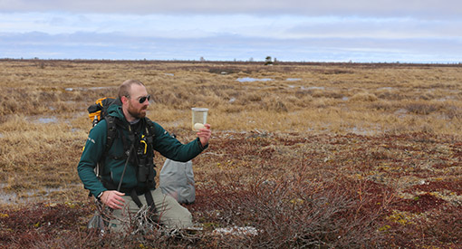 A Parks Canada employee kneels in the grass while holding a large glass of water with an egg in it.