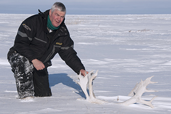 A man kneels in snow next to caribou antlers.