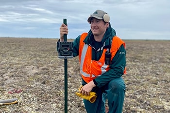 A Parks Canada employee kneels in the grass while holding a camera fastened to a stick.