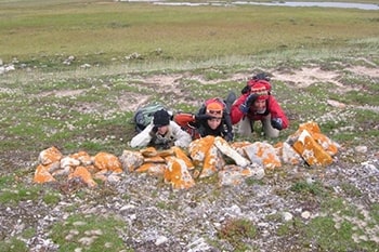 Rocks arranged in a line. Behind them, three people lie on their stomachs and face the camera.