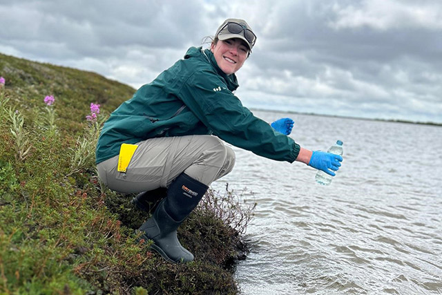 Une femme accroupie le long d’une colline tient une bouteille d’eau en plastique au-dessus d’un plan d’eau.