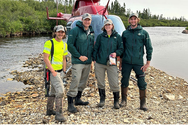 Four Parks Canada employees in uniform stand in front of a helicopter on rocky land with water on either side of them.