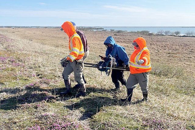 Quatre personnes transportent une grande pièce d’équipement de forage à travers un champ herbeux.
