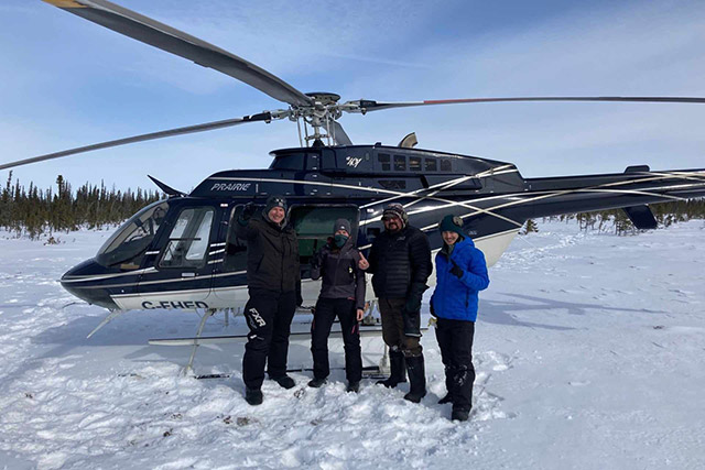 Four people in winter clothing stand in the snow in front of a helicopter.