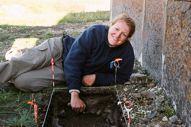 A person digs in a square hole while smiling and looking at the camera at Prince of Wales Fort National Historic Site.