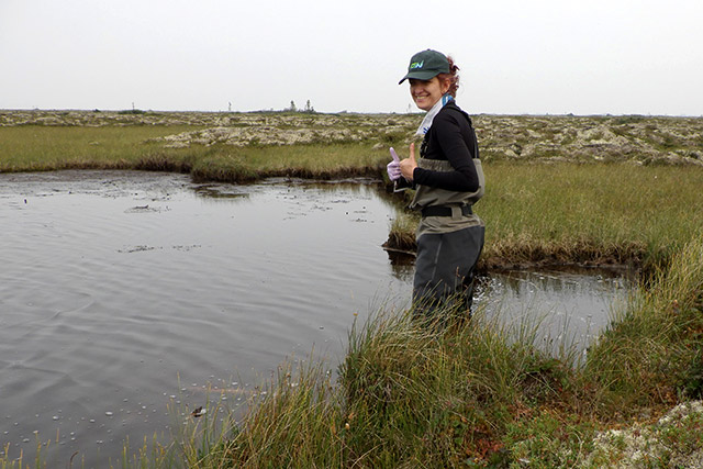 Une personne se tient près d’un étang et lève le pouce en direction de la caméra. Il y a de l’herbe et des rochers en arrière-plan.