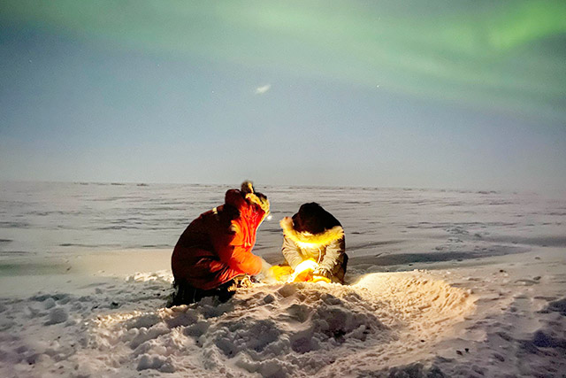 Deux personnes agenouillées dans la neige en projetant une lumière, avec des aurores boréales bien visibles dans le ciel.