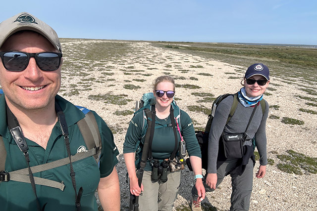 Trois personnes en uniforme de Parcs Canada marchent sur un terrain rocailleux et plat. 