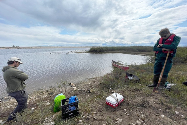 Deux personnes se tiennent près de la berge d’une rivière. Il y a une caisse et un étui d’équipement scientifique au sol.