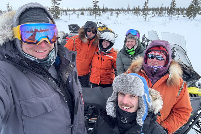 Sept personnes en vêtements d’hiver prennent un égoportrait avec des motoneiges et un paysage enneigé en arrière-plan. 