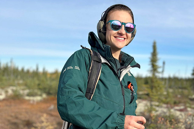 A Parks Canada employee in uniform smiles in a field with trees in the background.