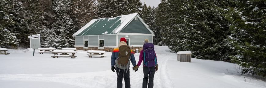 Two adults with backpacks walking in the snow on their way to the Point Wolfe shelter nestled in the snow covered forest