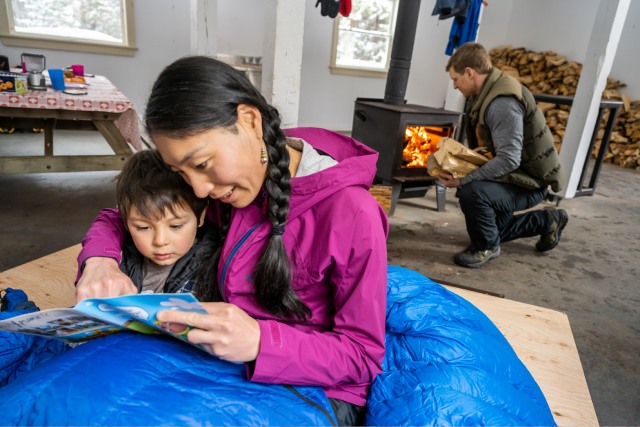 Soignant et enfant ensemble dans un sac de couchage bleu en train de lire un livre, tandis que le père ajoute du bois au feu à l'intérieur du poêle à bois
