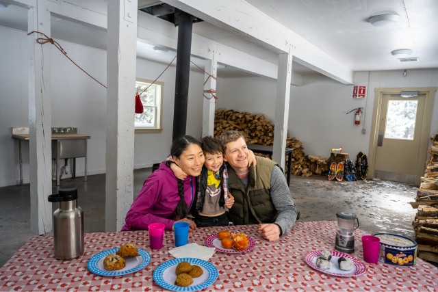 Caregivers and their child sitting closely at a picnic table surrounded by snacks. Stacked firewood is drying next to the wood stove in the background