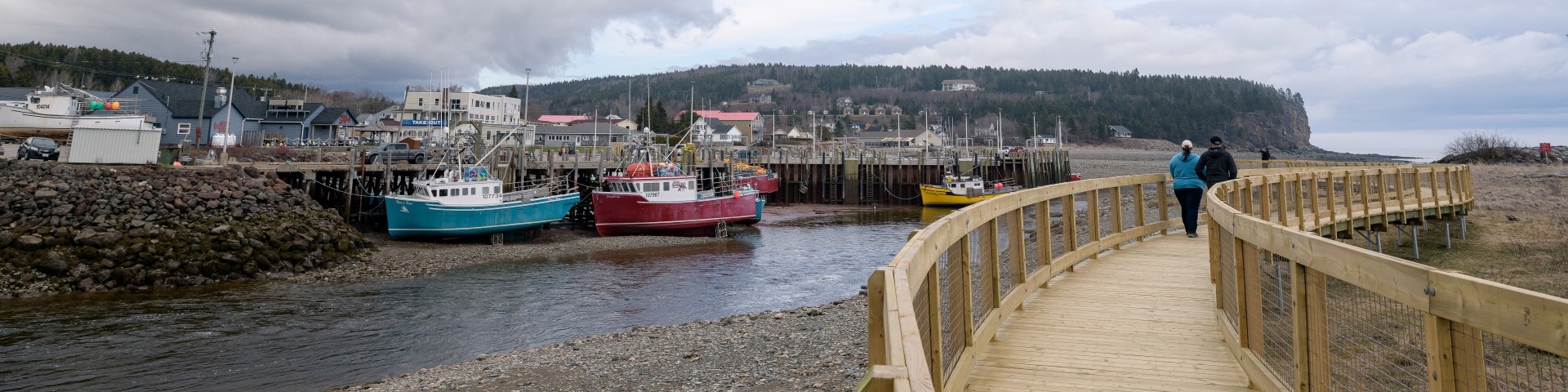 People walking on a boardwalk along the Upper Salmon River with fishing boats resting on the ocean floor at low tide.