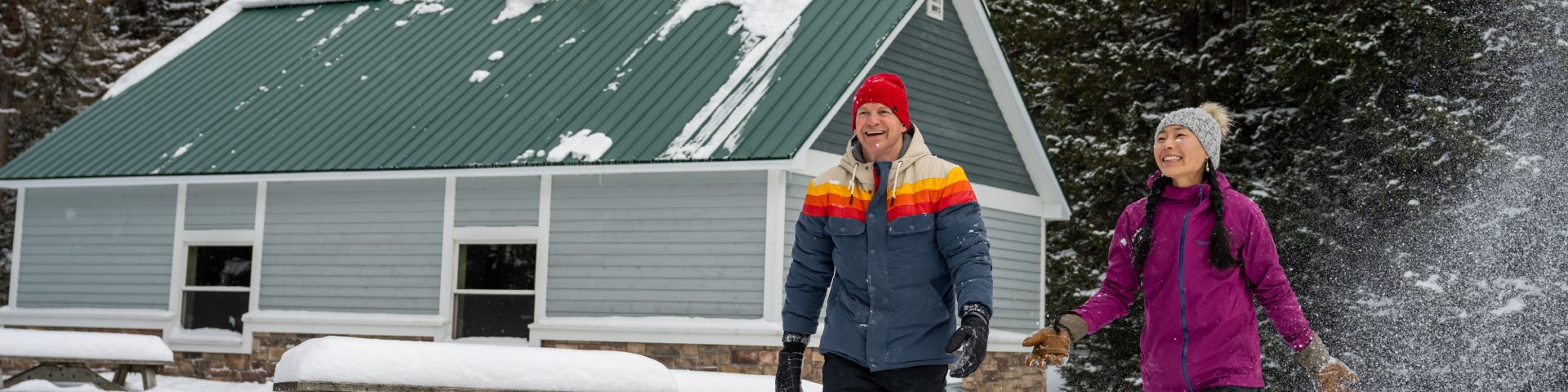 Deux adultes avec de grands sourires se promènent dehors avec les arbres et les tables de pique-nique recouverts de neige devant l'abri de Point Wolfe