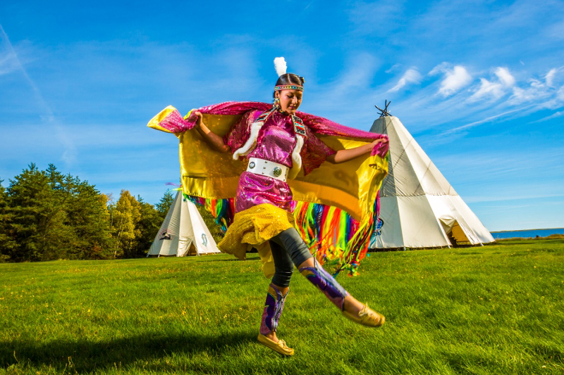 an Indigenous dancer wearing traditional regalia during a Mi'kmaw ceremony near the wigwams.