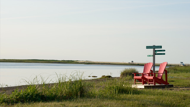 Deux chaises rouges au bord de l'eau, avec un panneau indiquant 'Kouchibouguac'.