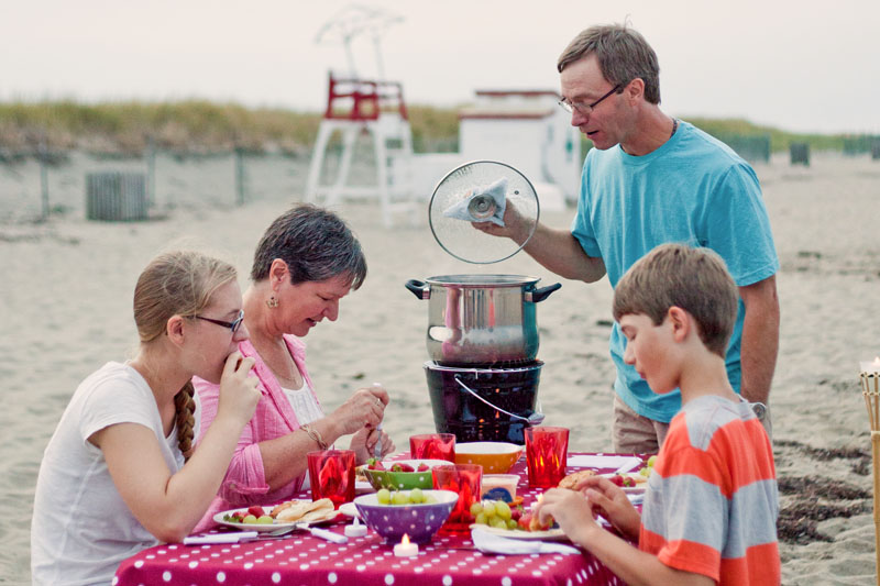 Une famille mange un pique-nique sur la plage alors que le père cuit des coques.