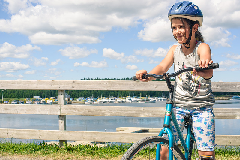 A young girl on her bike near a wooden fence, with a body of water in the background.