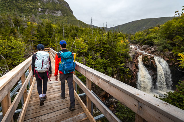 Two people walking across a bridge admiring a waterfall on the Gros Morne Mountain approach trail
