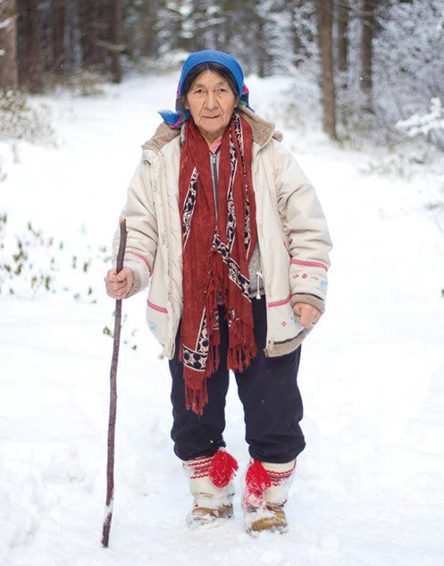 Une femme autochtone face à la caméra, avec une canne, dans la neige.