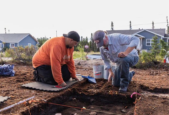 Deux personnes agenouillées travaillant ensemble dans un champ. Elles creusent et examinent le sol. Des maisons et des arbres sont visibles à l'arrière-plan.