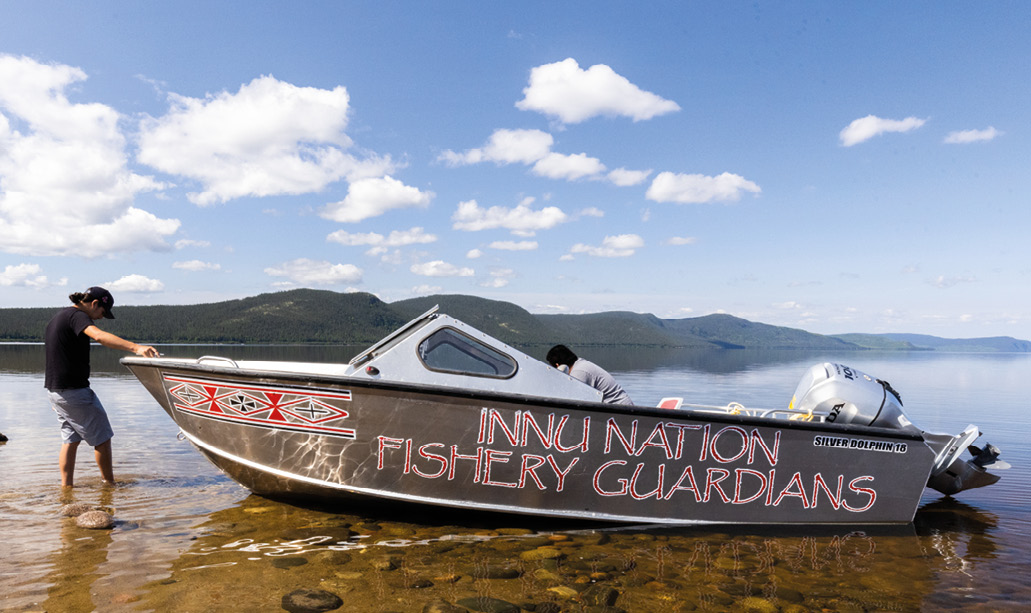 Deux personnes, l'une à l'intérieur et l'autre à l'extérieur d'un bateau des Gardiens des pêches de la Nation innue. Eau et ciel bleu en arrière-plan.
