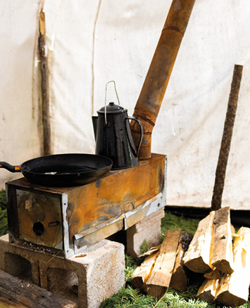 Une cuisinière avec une bouilloire et une poêle à frire, située à l'intérieur d'une tente, avec du bois empilé à côté.
