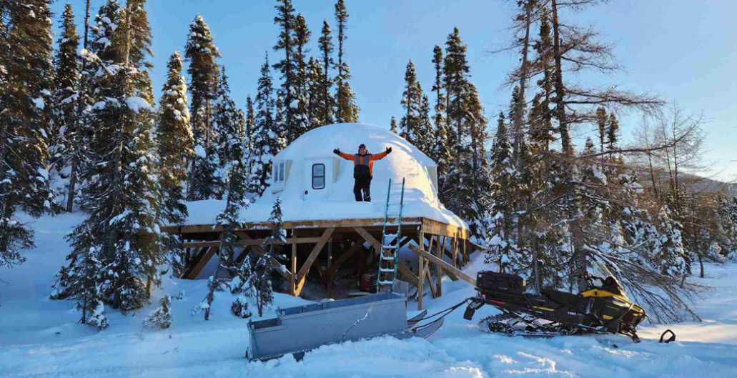 Une personne debout au sommet d'une cabane recouverte de neige, avec une motoneige en hiver.