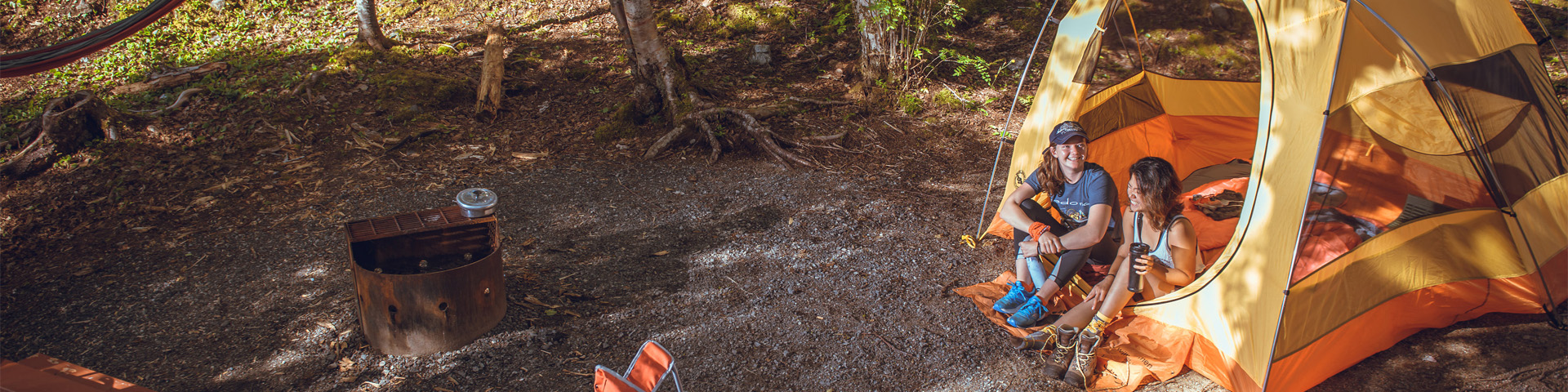 two people sit in a yellow tent next to a picnic table, orange camping chair and a fire pit