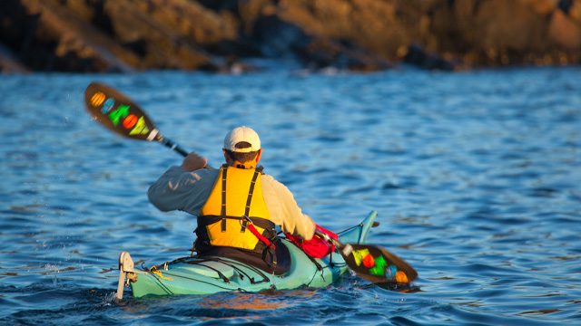 A kayaker paddling away from the camera with a brightly-coloured life jacket and paddles