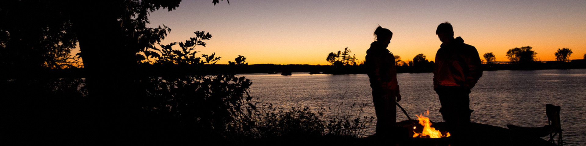 Two people in silhouette standing next to a coastal campfire at sunset.