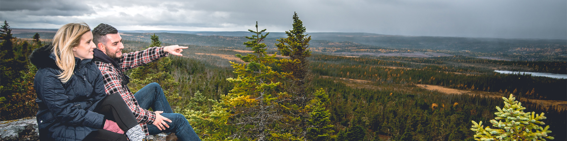 two people sitting on a rocky cliff, overlooking a forest park