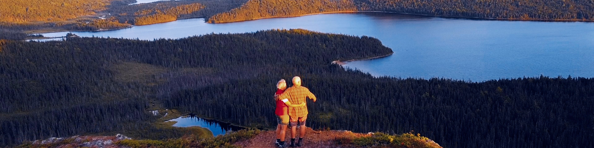 two people standing a cliff overlooking a pond surrounded by trees