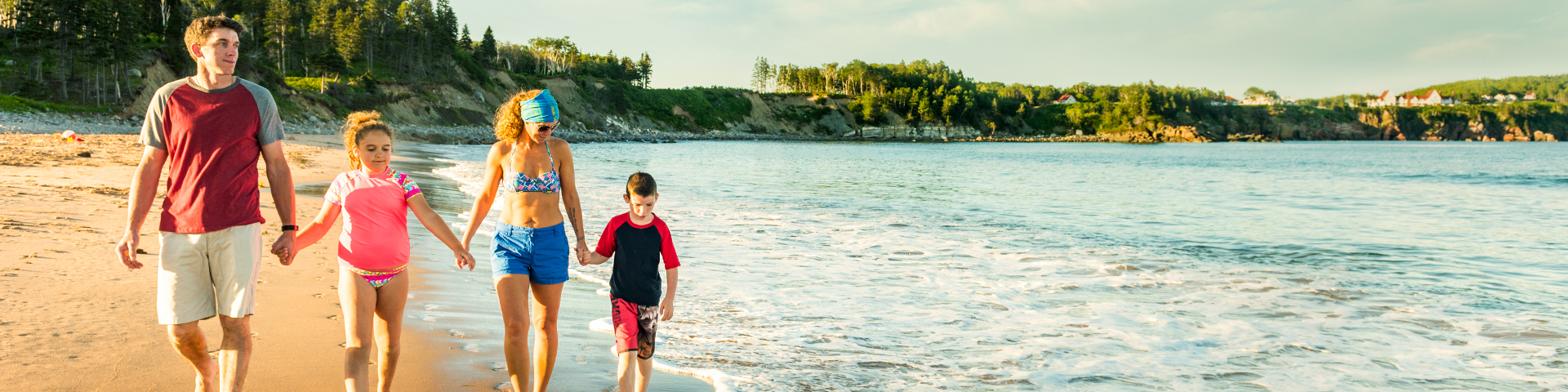 A family of four walks barefoot along a sandy shoreline as gentle waves lap at their feet.