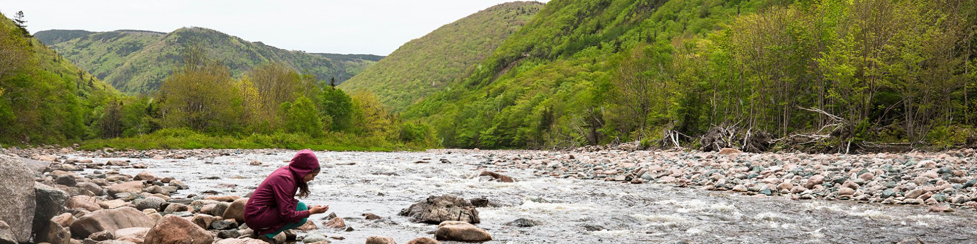 Child picking up a rock from the river with the green trees and hills in background.