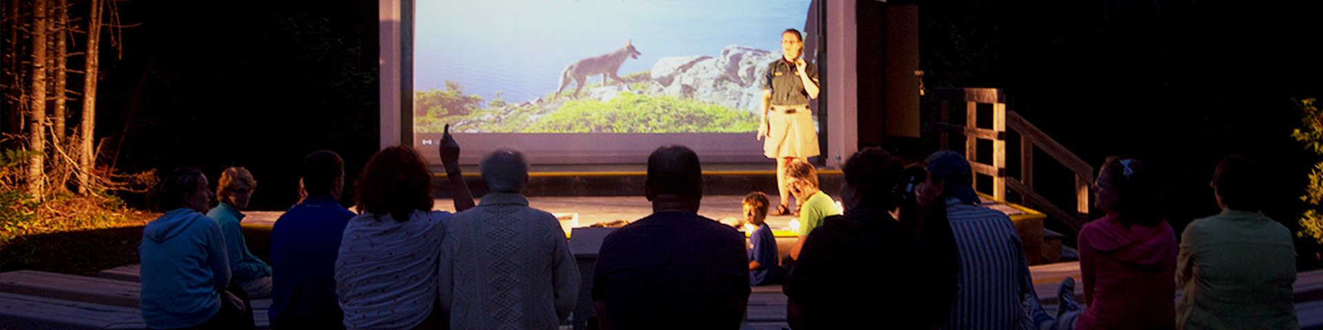 In an open-air theatre, visitors gather to hear an interpreter share a story, with a large screen illuminating the night.