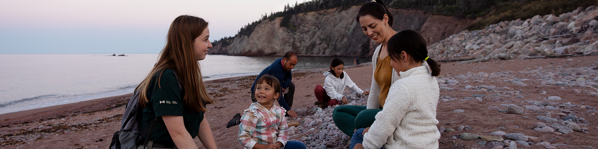 An interpreter sitting on the sand with a family of five, all gathered together on a beach and engaged in conversation near the shoreline.