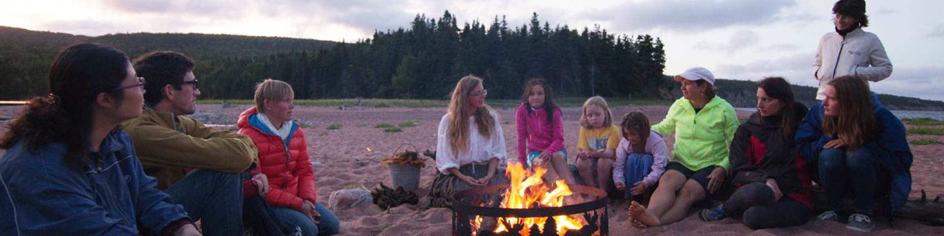 A group of people sitting around a campfire on a beach at dusk.