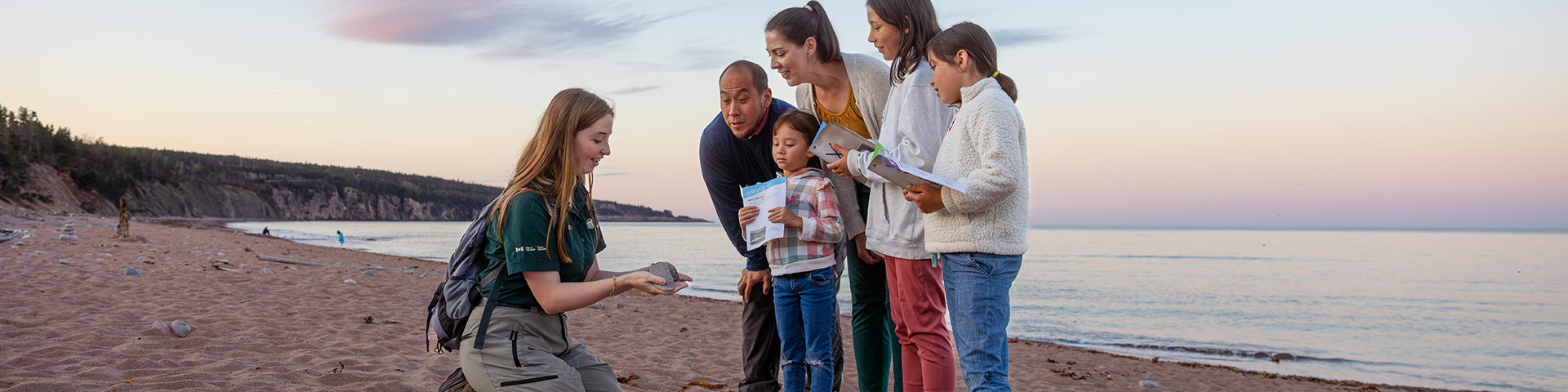 A family interact with a Parks Canada interpreter who is holding a shell on the beach. 