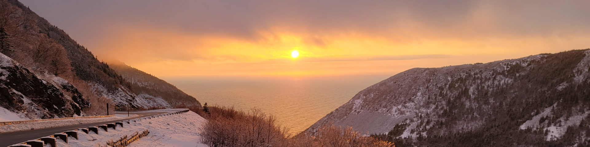 A winter's sunset overlooking the ocean and the Cabot Trail in Cape Breton Highlands National Park with an orange and pink sky in the background.  