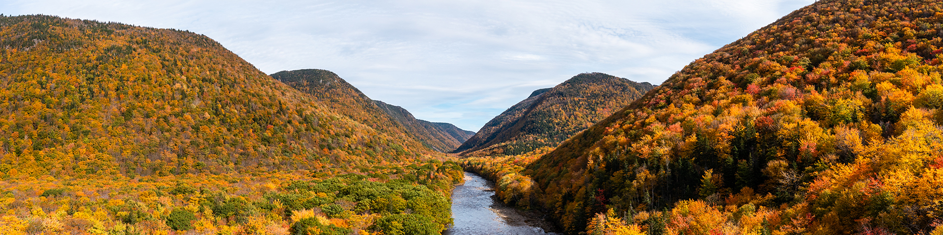 Une vallée fluviale entourée de feuilles d'automne vibrantes dans les tons rouge, jaune, orange et vert, bordant les rives et les collines.