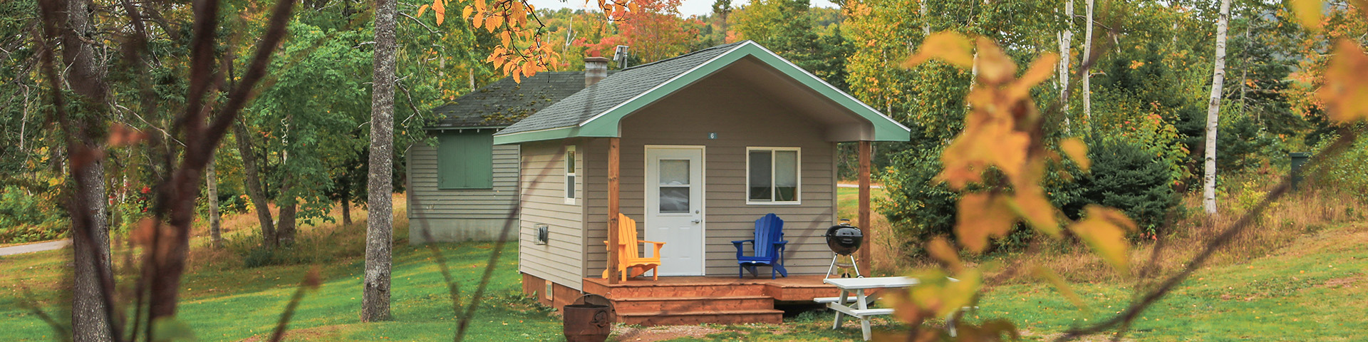 une image d’un chalet à pignon couverte entourée d’arbres verts et de feuillage jaune. La cabine a un porche couvert avec deux chaises et un barbecue au charbon.