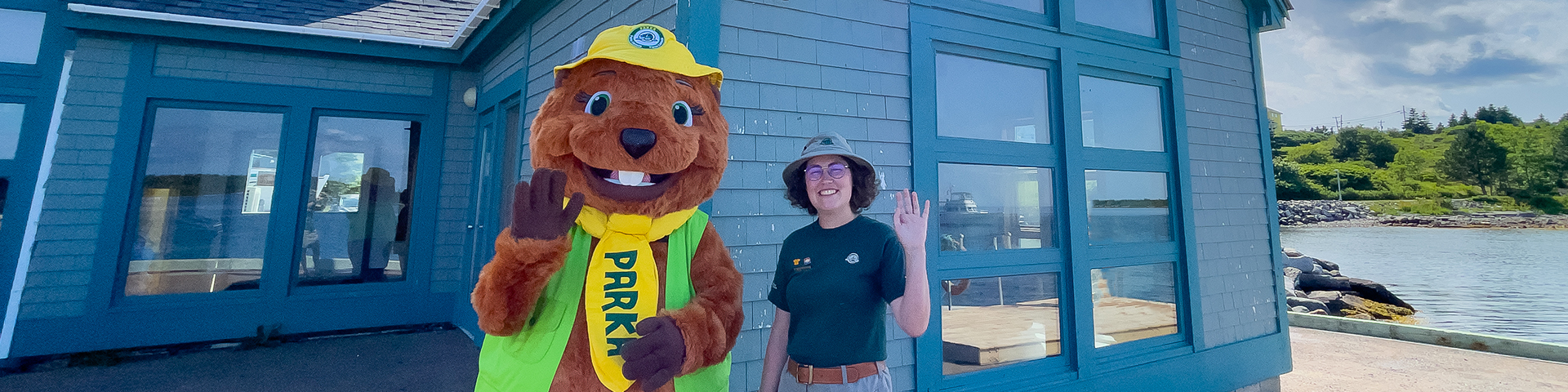 Une mascotte de Parcs Canada et un employé de Parcs Canada saluent joyeusement la caméra.