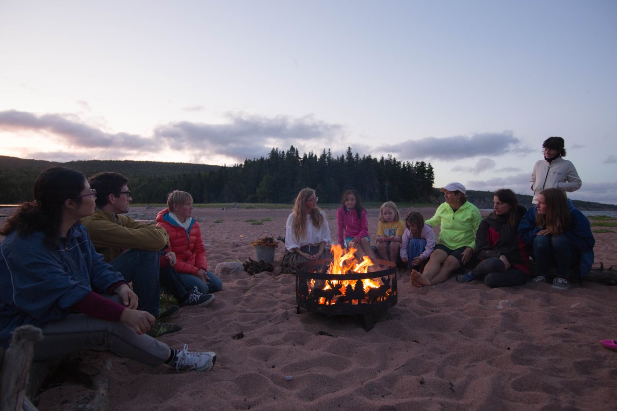 A group of people sitting around a campfire on a beach at dusk.