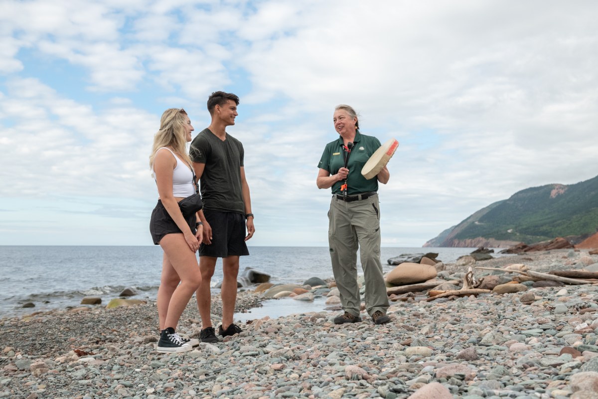A Parks Canada interpreter and two visitors stand on the rocky beach at Mkwesaqtuk/Cap-Rouge