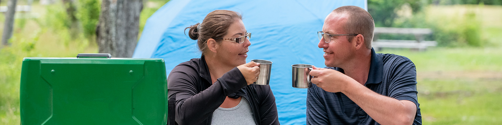 Un homme et une femme sont assis à côté d'un four de camping devant leur tente turquoise. Ils brandissent leurs tasses de camping et se sourient.