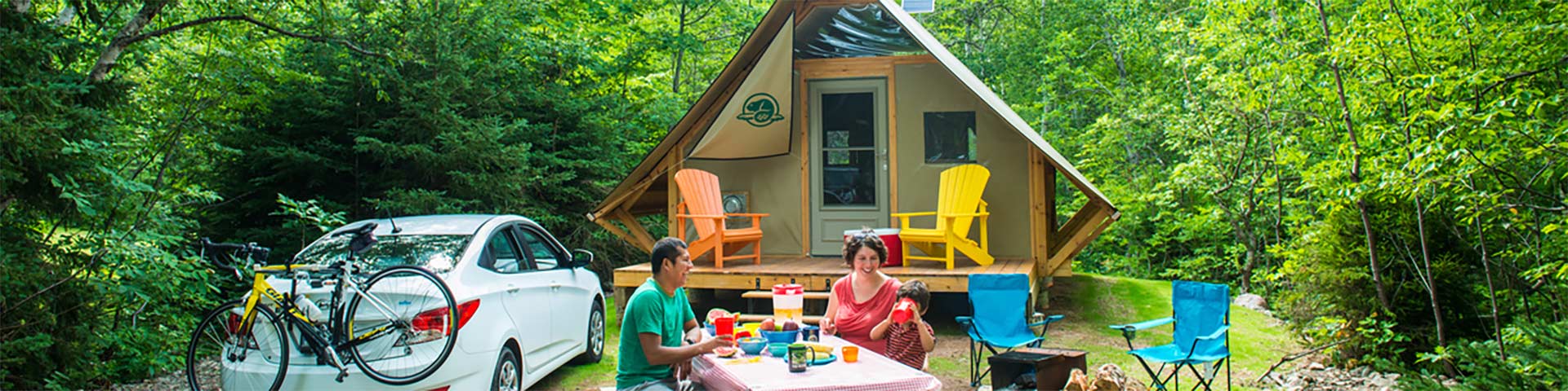 Two adults and a child sit at a picnic table in front of a Parks Canada Otentik, there is a parked car with a bicycle on the back to the left and a fire pit with two campaign chairs to the right.
