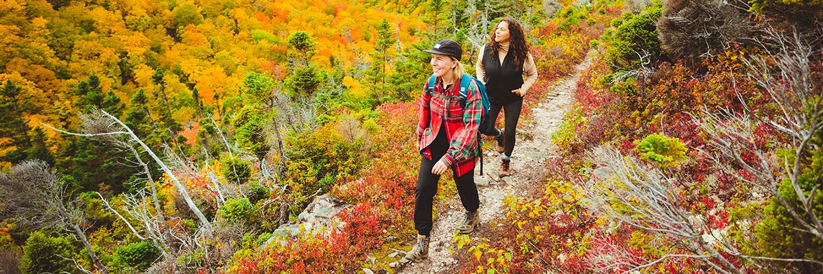 Two smiling women hike on a path surrounded by bright autumn leaves